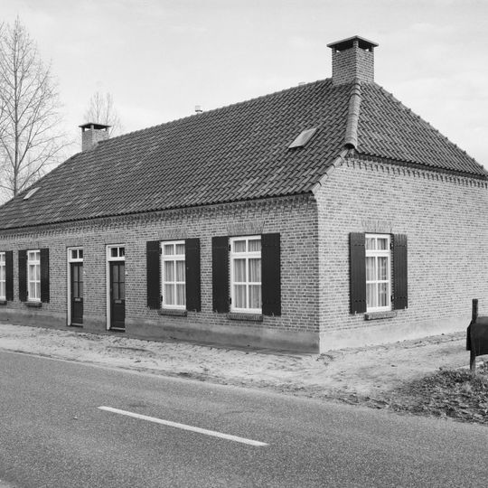 Dubbele woning. Dwarshuis onder met pannen gedekt wolfdak. Baksteenfries met zaagtandlijst. Vensters met luiken