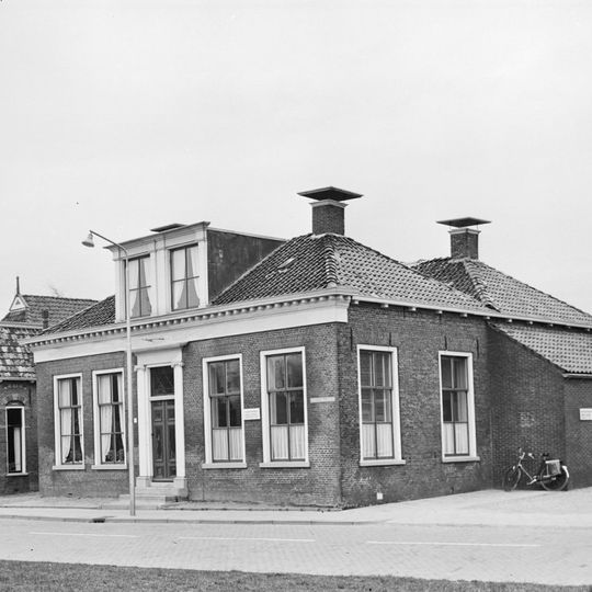 Laag woonhuis met voorste deel met omlijste ingang en geblokte kroonlijst onder laag schilddak met hoekschoorstenen waarop borden en waarschijnlijk jongere dakkapel