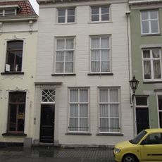 Huis met gebosseerd gepleisterde lijstgevel, kroonlijst op klossen en ingang tussen gecanneleerde pilasters. Plat dak
