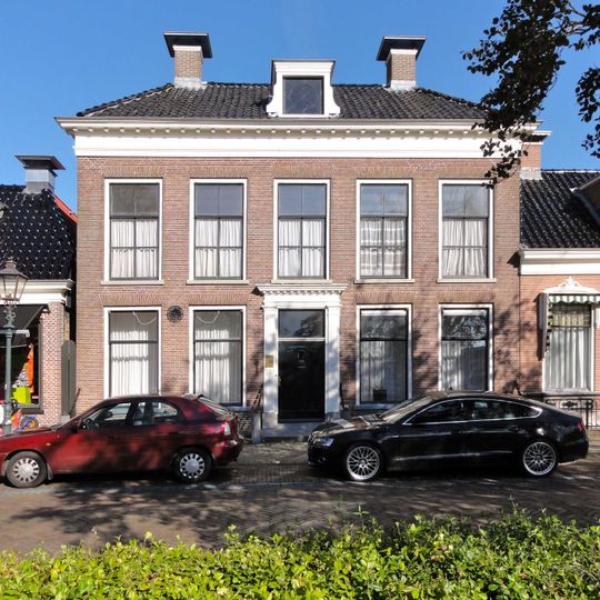 Vijf venstertraveeën breed woonhuis met omlijste ingang en verdieping onder laag schilddak met dakkapel en hoekschoorstenen waarop borden