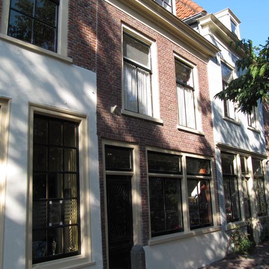 Pand met verdieping onder schilddak. Lijstgevel waarin boven twee T-vensters. Deur met gietijzeren roostervulling en medaillon