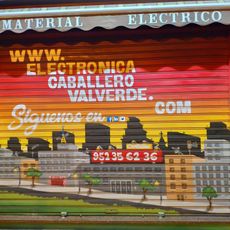 Mural en la persiana de cierre de un establecimiento comercial que presenta un paisaje urbano en tonos anaranjados en el que se ven varios edificios de apartamentos.