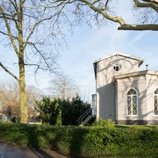 Gepleisterd 6-hoekig gebouwtje met spitsboogvensters, waarin authentieke ruitvormige roedeverdeling. Fijn gedetailleerde kroonlijst met consoles